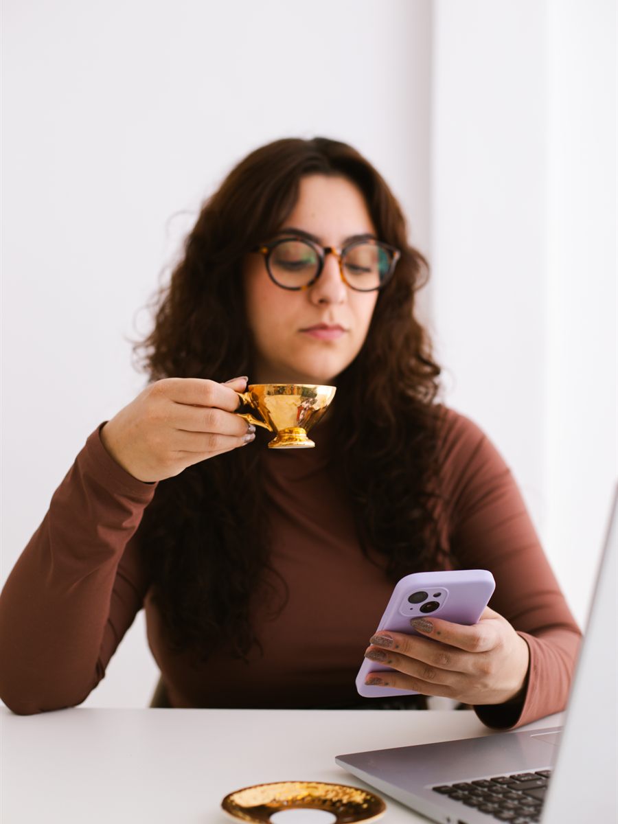 Ana Vitória tomando café na mesa de trabalho, com o celular e o notebook
